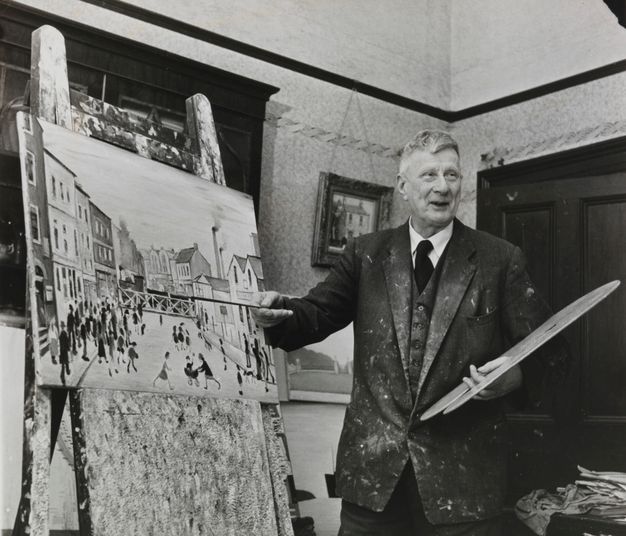 A black-and-white photograph of an older artist in a suit, holding a paintbrush and standing in front of an easel displaying a cityscape painting.