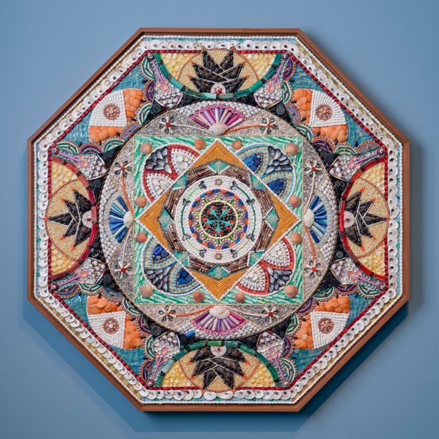 A vibrant octagonal shellwork piece with a geometric mandala-like design, combining orange, blue, and green elements.