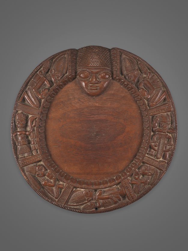 A circular wooden plaque with elaborate carvings of human and animal figures around the border, featuring a prominent human face at the top centre.