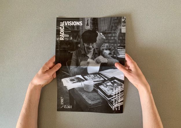 Two hands holding a black-and-white magazine titled "Radical Visions," featuring a cover photograph of a person wearing a hat and glasses while working with papers at a cluttered desk. The desk contains several magazines and books, and a cup with a distinct zigzag pattern. The text "1972/1987" appears on the cover's left side, alongside additional text at the bottom. The image conveys themes of journalistic and creative endeavors during the mentioned period.